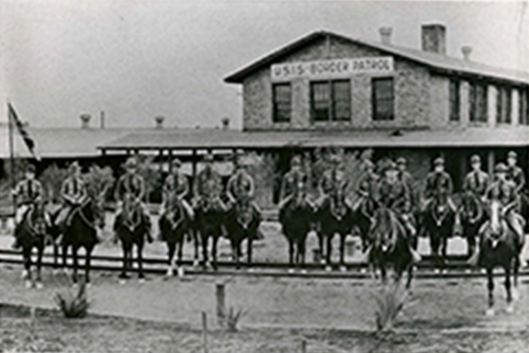 Building in the background has border patrol sign with men on horses waiting in front of the building