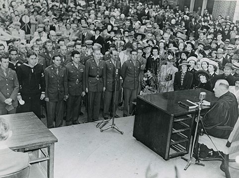 Man sitting at a large desk in front of large room with many people standing to take the oath.