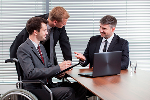 Man in a wheelchair sitting at a large table talking with two other men while looking at a laptop monitor