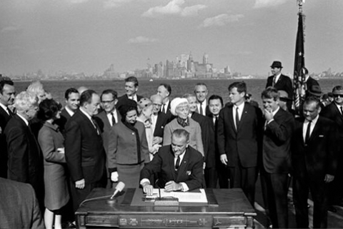 Image of President Lyndon B. Johnson signs the Immigration and Nationality Act as Vice President Hubert Humphrey, Lady Bird Johnson, Muriel Humphrey, Sen. Edward (Ted) Kennedy, Sen. Robert F. Kennedy, and others look on. *LBJ Presidential Library.