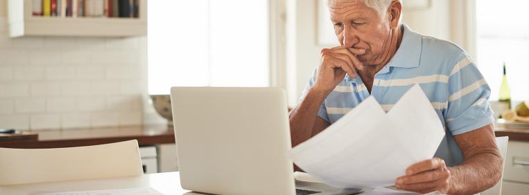 Older man sitting at a table looks down at a computer screen and is holding papers in his other hand